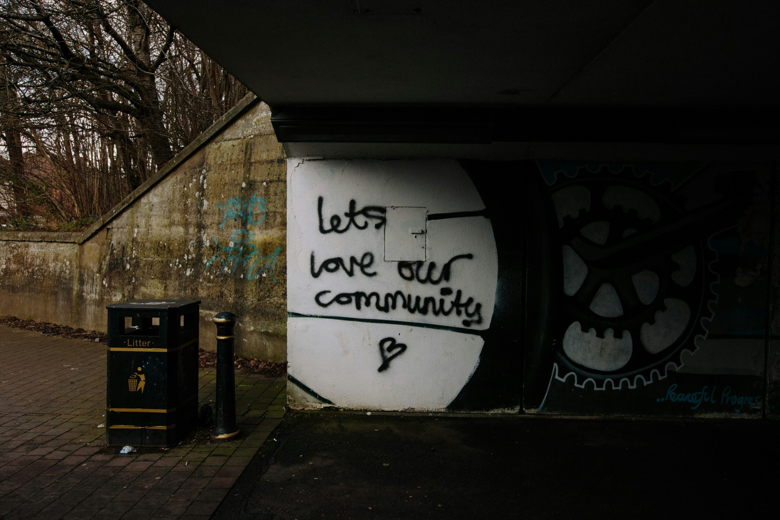 Graffiti reading "let's love our community" beneath a dark overpass, reflecting the themes of inheritance, identity, and the Light Omens factions.